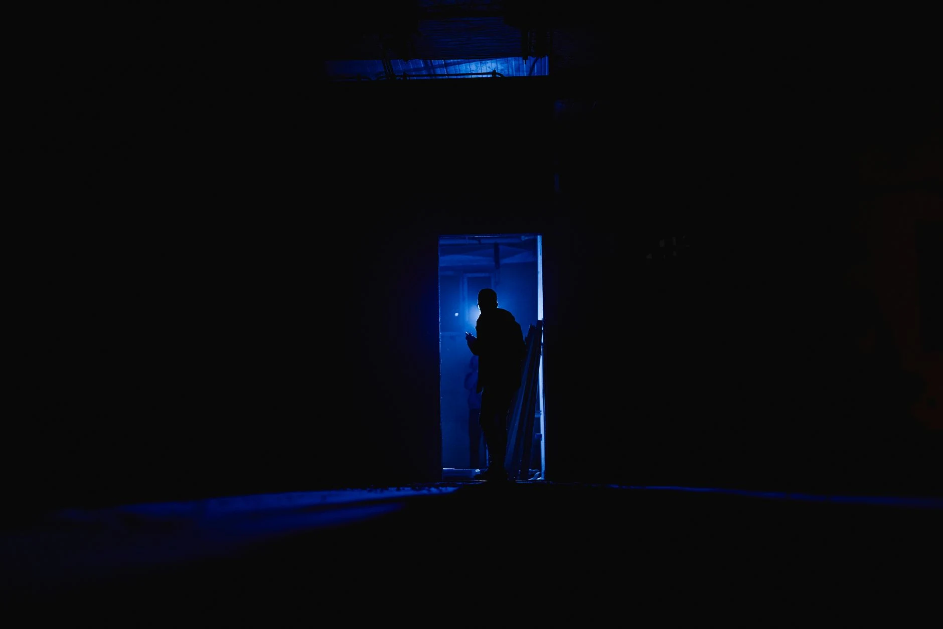 A mysterious silhouette stands in a dimly lit room, bathed in blue light, suggesting intrigue and solitude.