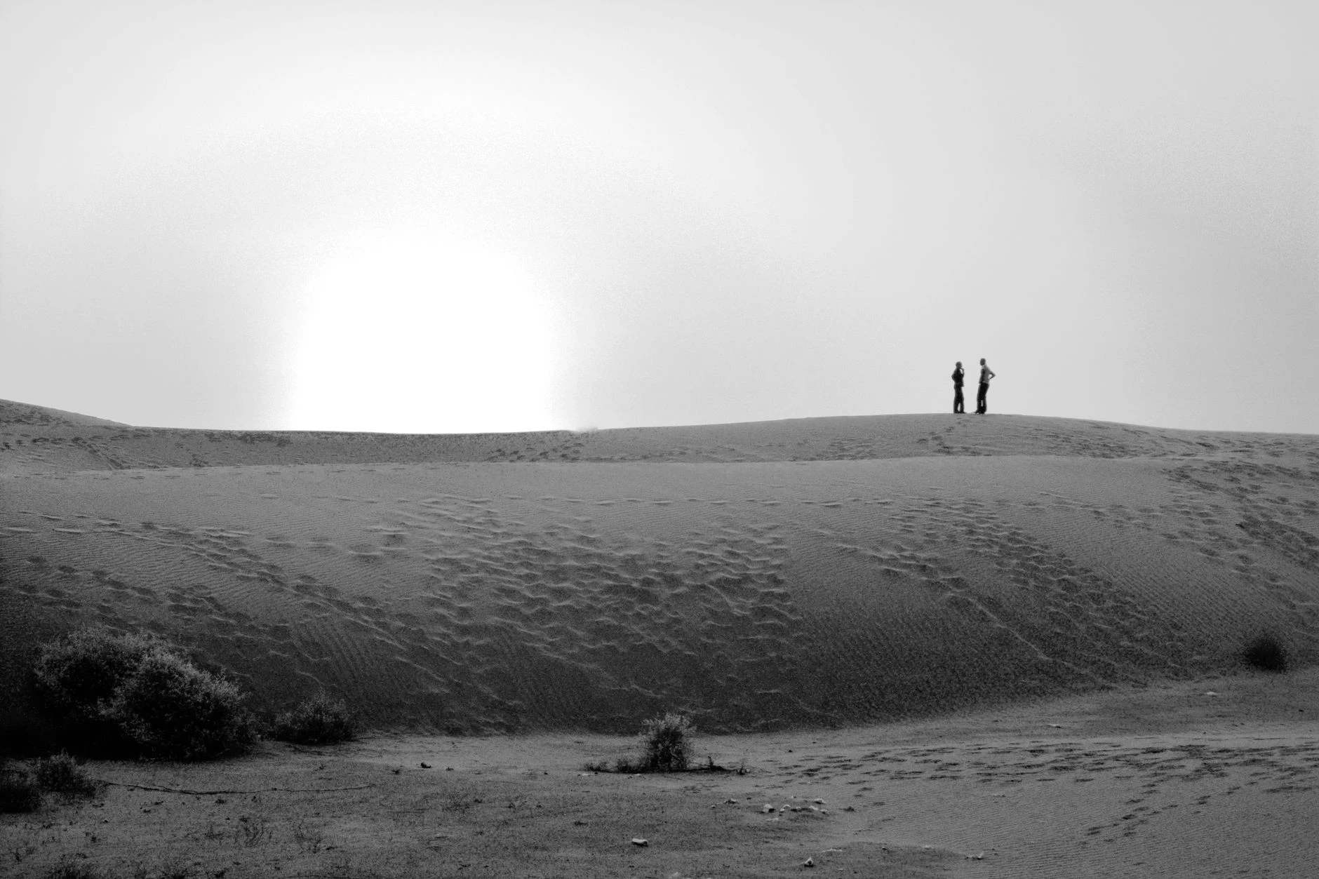 Black and white photo of two people standing on a sand dune, silhouetted against the sunset.