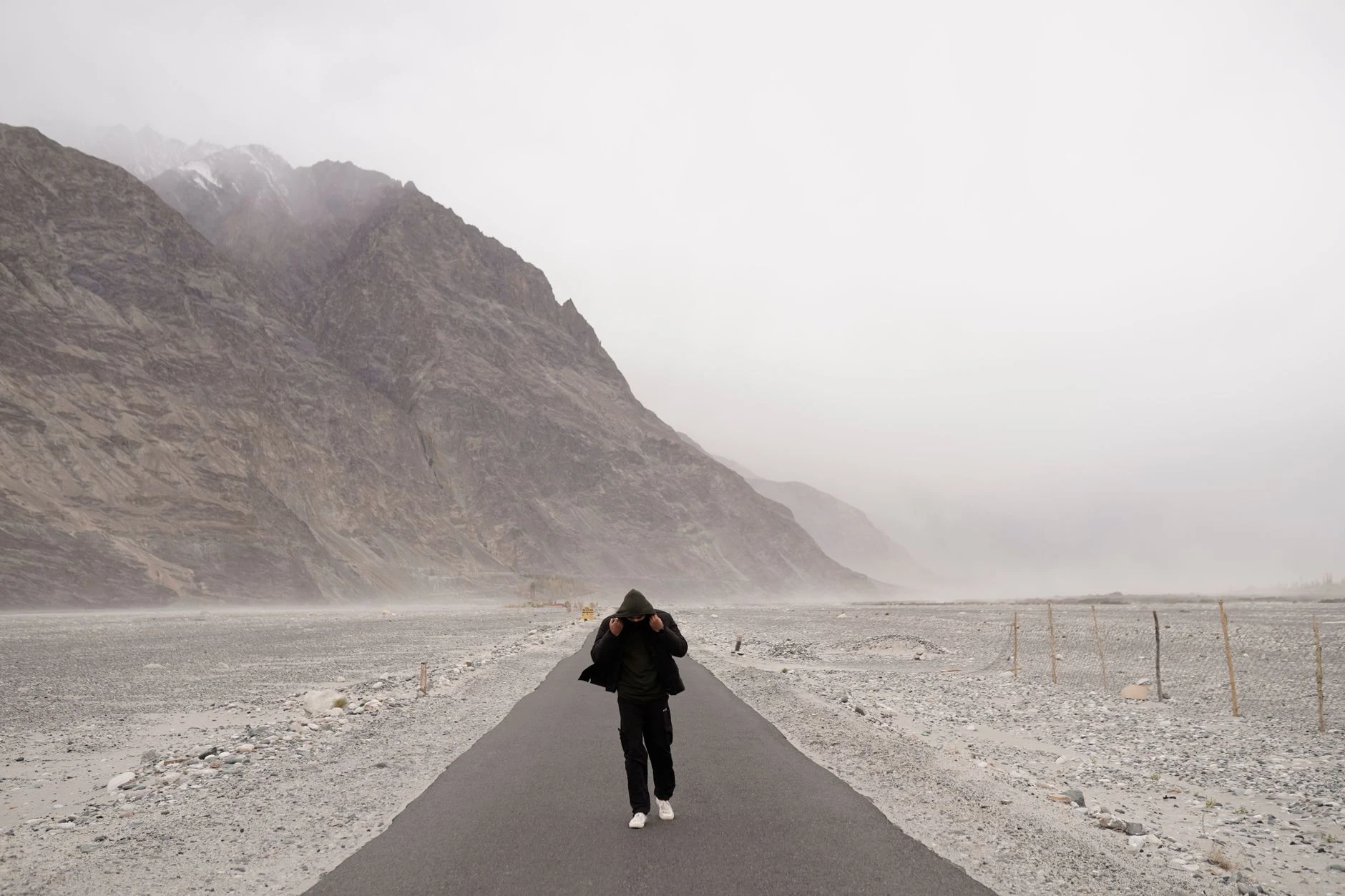 A solitary person walks on a road through a rugged, mountainous desert under an overcast sky.