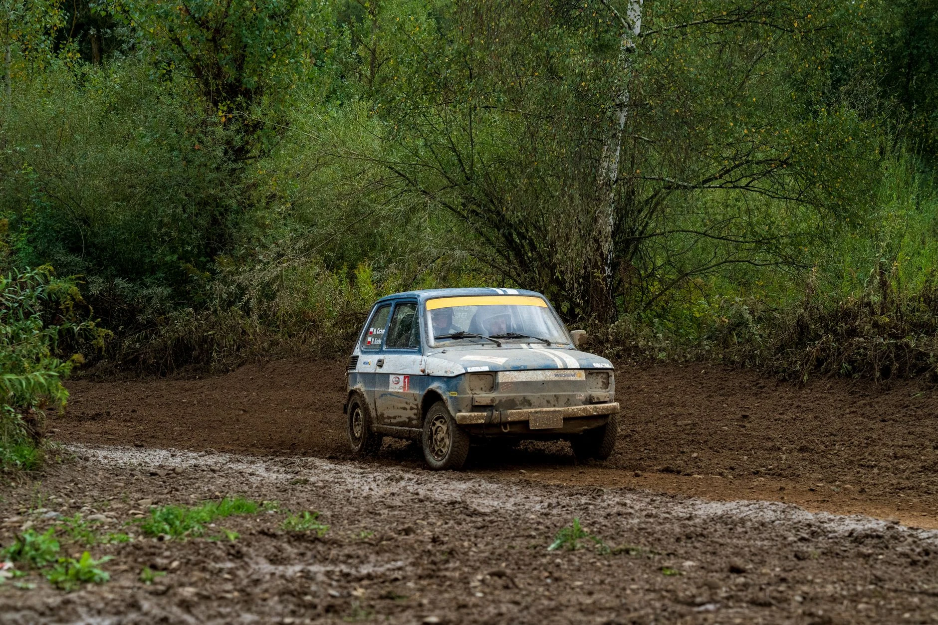A vintage car navigates through a muddy and forested rally track in Poland.